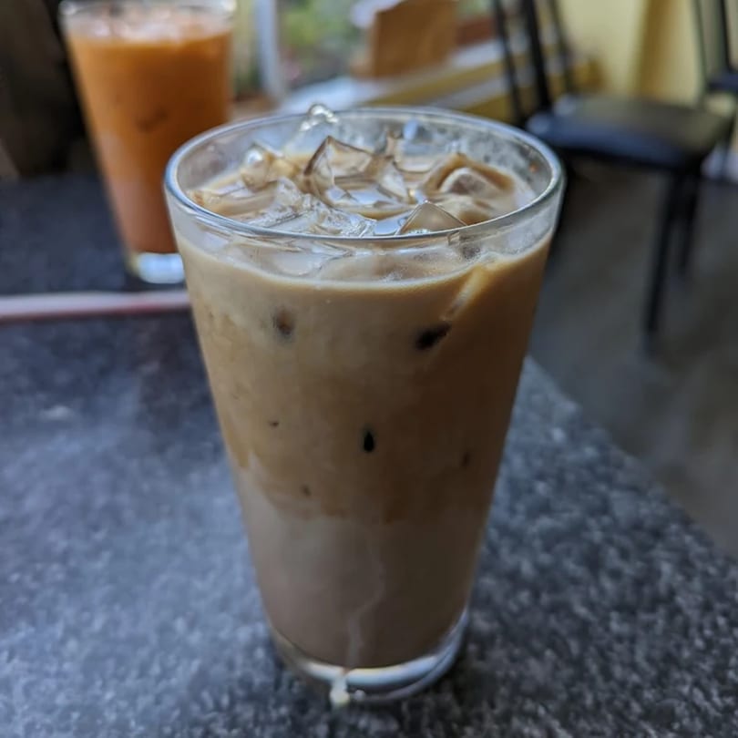 Iced Thai coffee with ice cubes in a glass, with another drink blurred in the background.