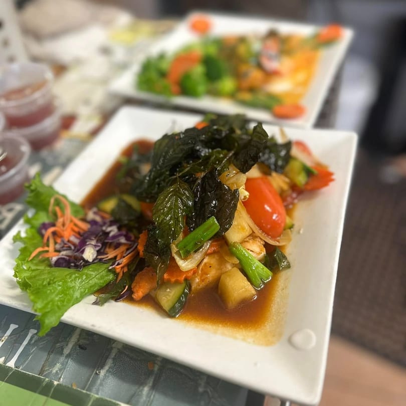 A Thai stir-fry with dark leafy greens, vegetables, and a brown sauce on a white plate.